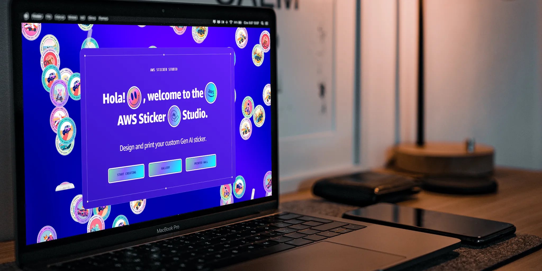 A laptop showcasing the AWS Sticker Studio interface on a wooden desk, featuring a welcome message in Spanish and colorful stickers, alongside a smartphone and notebook, all illuminated by a stylish lamp.