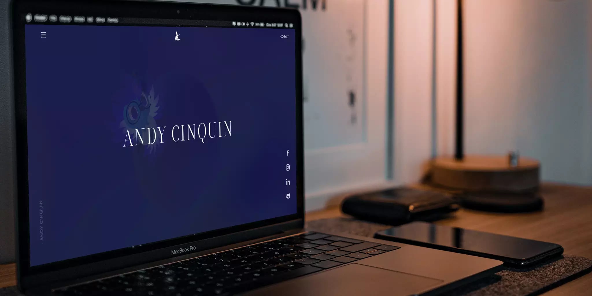 Close-up of a laptop displaying 'ANDY CINQUIN' name, social media icons for Facebook, Instagram, and LinkedIn, smartphone, and lamp on a desk in a warmly lit home or office setting