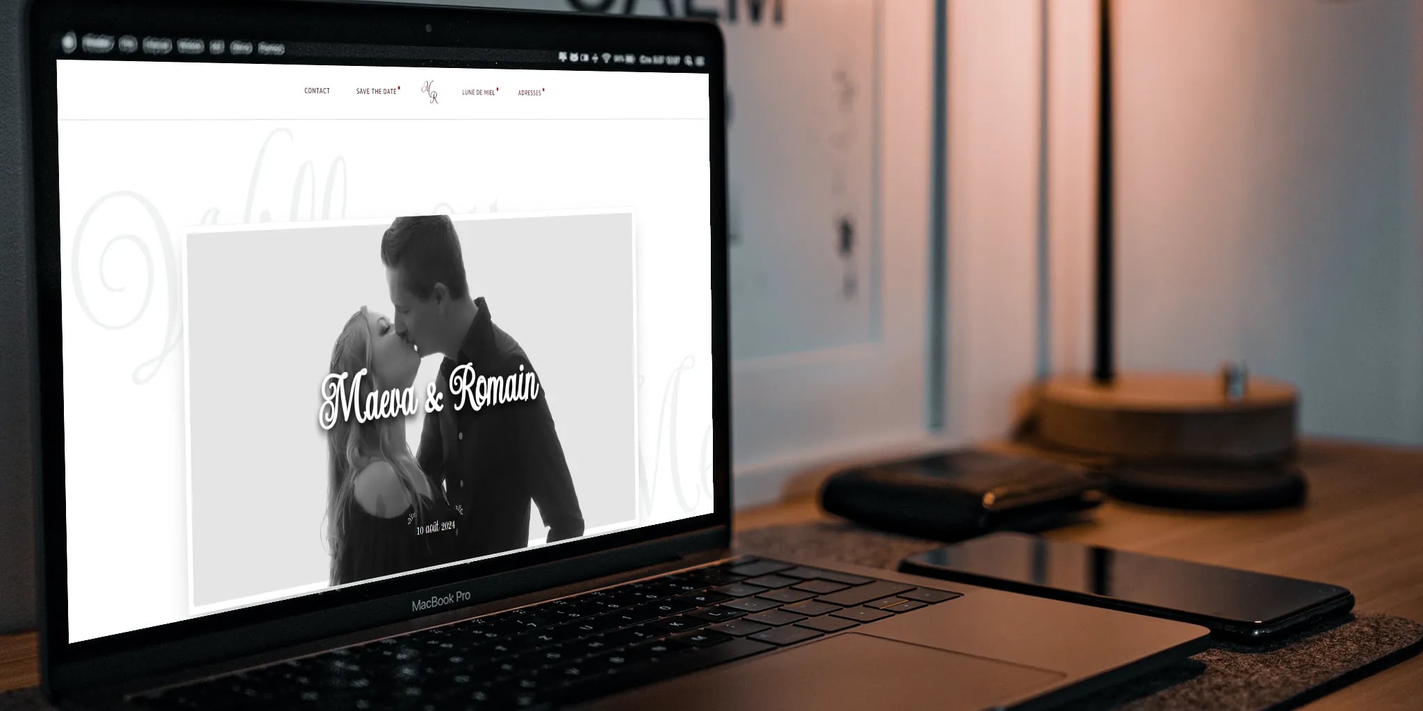 Laptop displaying a wedding website with a couple embracing photo, smartphone, and desk lamp on a desk