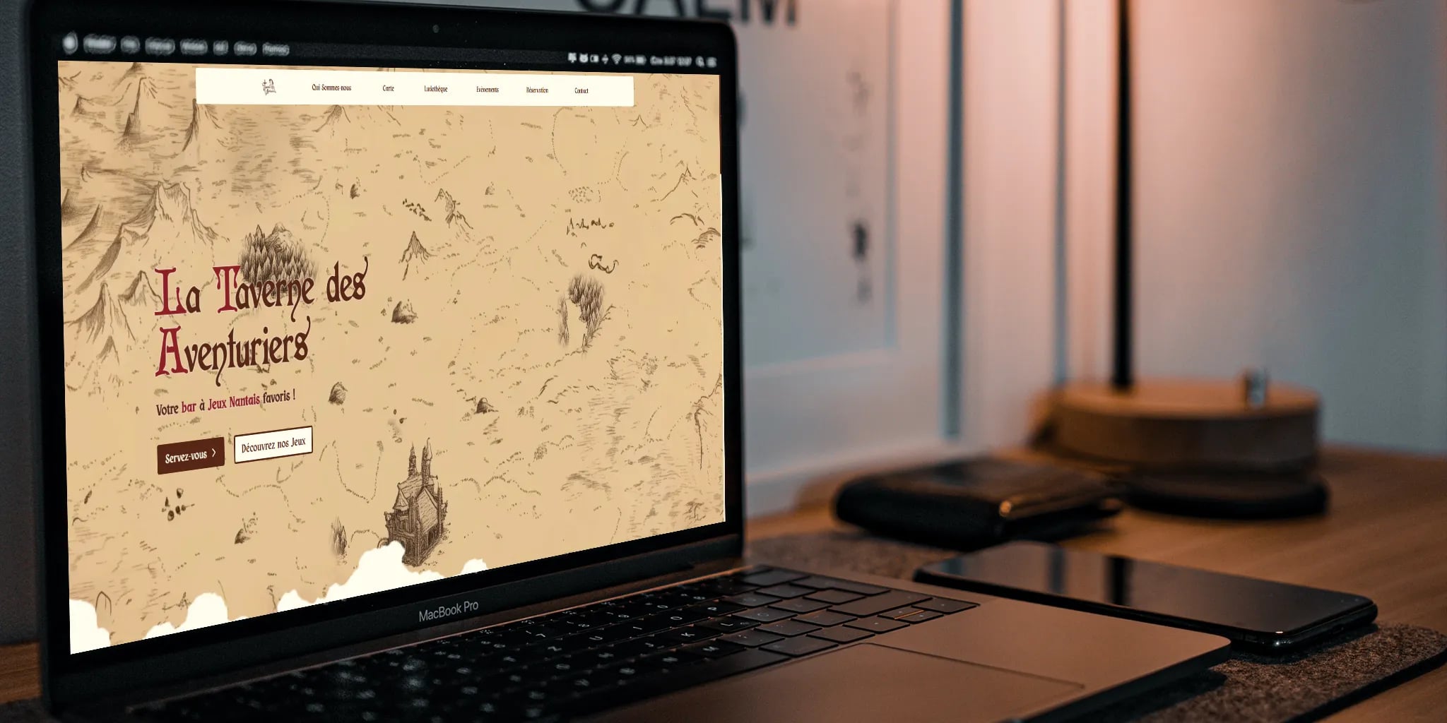 A cozy workspace with a laptop displaying the La Taverne des Aventuriers website, featuring bold red text and surrounded by a smartphone and a closed notebook on a wooden desk.