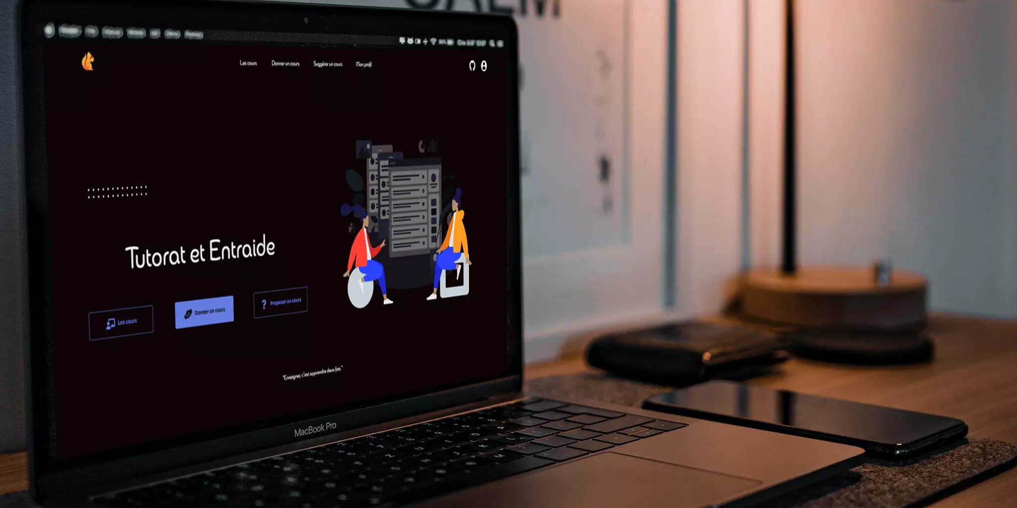 Laptop displaying Tutorat et Entraide website with two illustrated characters and smartphone on desk